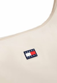 Bolso beige con una textura suave, que presenta un parche de marca cosido en azul marino y rojo etiquetado como "TOMMY JEANS". Cierre en la parte superior.