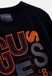 Musta collegepaita, jossa on monivärinen kirjailtu teksti oranssina ja sinisenä, pyöreä kaula-aukko ja näkyvät sauma-yksityiskohdat.