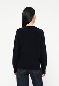 Mujer con cabello largo y negro, vestida con un suéter azul marino oscuro y jeans azul oscuro, de espaldas contra un fondo blanco liso.