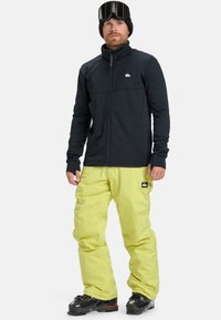 Zwarte ritsjack met hoge kraag, gecombineerd met losse, felgele skibroek. Draagt een zwarte beanie en skibril, staand rechtop.