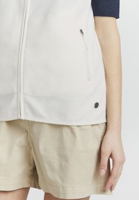 Weißer Fleece-Weste mit einem Vorderreißverschluss, Seitentaschen und einem schwarzen Logo-Akzent nahe dem Saum. Getragen über beigen Shorts.