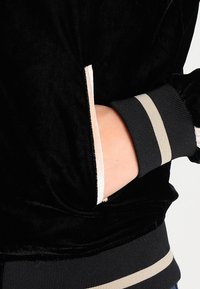 Giacca di velluto nero con polsini a strisce grigie e crema, dotata di tasca laterale. Tessuto morbido e dettagli di design casual vicino al polso.