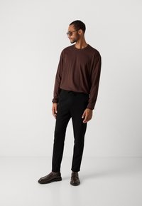 Hombre con camisa de manga larga marrón, pantalones negros, mocasines marrones y gafas de sol, de pie contra un fondo claro y liso.