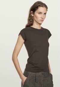 T-shirt aderente marrone scuro con maniche corte, scollatura rotonda e dettagli di cucitura sottili, abbinato a pantaloni grigi testurizzati.