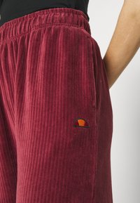 Bordeaux kordflorbyxor med resårmidja, med vertikal ribbad textur och en liten broderad logotyp nära vänster höft.