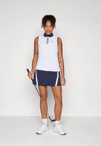 Γυναίκα που στέκεται με ένα κλαμπ γκολφ, φορώντας ένα αμάνικο λευκό πόλο μπλουζάκι και ναυτικό μπλε φούστα με λευκές λεπτομέρειες και λευκά αθλητικά παπούτσια.