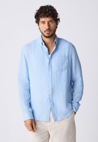 Helesinine linane pikkade varrukatega nööbitav särk, millel on rinnatasku ja peen tekstuur. Sobitatud helevärviliste pükstega.