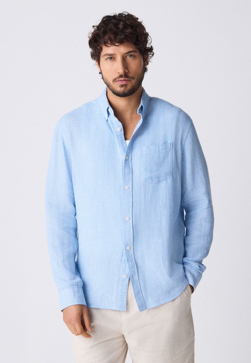 Helesinine linane pikkade varrukatega nööbitav särk, millel on rinnatasku ja peen tekstuur. Sobitatud helevärviliste pükstega.