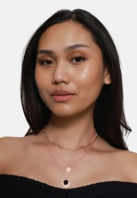 Trois colliers superposés avec des chaînes en or rose, un pendentif circulaire avec une pierre noire et un charm décoratif en forme de soleil.