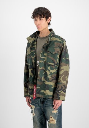 Young man wearing green camouflage jacket with red "Remove Before Flight" tag and ripped blue jeans, standing against white background.