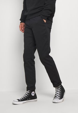 Person trägt schwarze lässige Hose, schwarze High-Top Converse-Sneaker und ein schwarzes Langarmshirt, steht mit den Händen in den Taschen.
