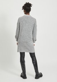Maglione grigio oversize con maniche lunghe, lavorato a maglia con texture, e orlo asimmetrico, indossato sopra leggings in pelle nera e stivali neri al ginocchio.