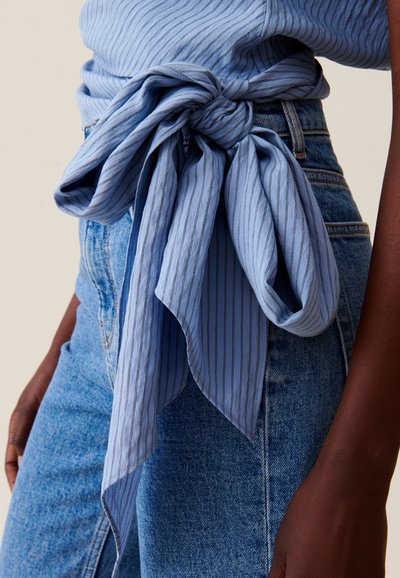 Top en tissu bleu clair à rayures verticales avec un nœud à la taille, associé à un jean en denim bleu délavé taille haute. Texture lisse.