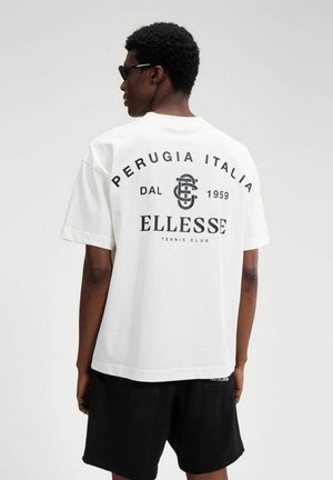 Osoba z krótkimi kręconymi włosami w okularach przeciwsłonecznych, biała koszulka z czarnym napisem „Ellesse Tennis Club” i czarne szorty, stojąca tyłem.