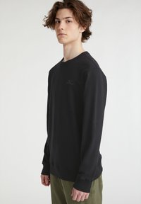 Sudadera negra de manga larga con cuello redondo, hecha de un material suave. Presenta un logo sutil y puños acanalados. Combinada con pantalones verde olivo.
