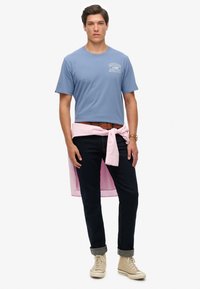 Camiseta azul con estampado de "SUPERDRY ATHLETICS", camisa rosa atada a la cintura, jeans oscuros y zapatillas ligeras color beige. Vestimenta casual.