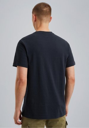 Man met kort haar die een effen donkerblauw t-shirt met korte mouwen en olijfgroene cargoshorts draagt, staand tegen een lichtgrijze achtergrond.