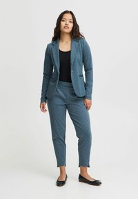 Türkiser Anzug aus strukturiertem Material, bestehend aus einem taillierten Blazer mit Reverskragen und Punkten. Die Hose ist schmal zulaufend und hat seitliche Schlitze.