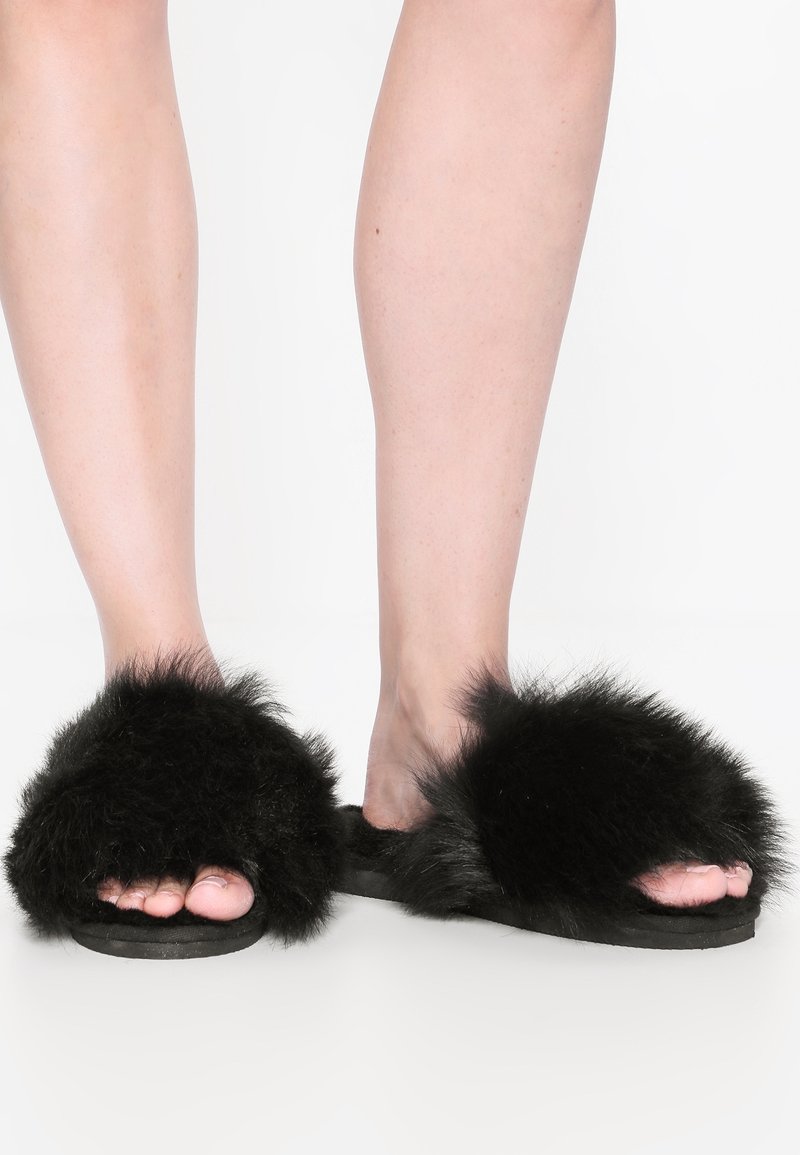 Pieds portant des pantoufles noires en fourrure à bouts ouverts, posés sur une surface blanche sur un fond uni.