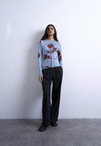 Femme debout contre un mur blanc, portant une chemise à manches longues bleu clair avec un imprimé floral rouge, un pantalon large noir et des chaussures noires.
