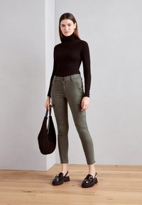 Črn rebrast pulover s turtleneck izrezom, olivno zelene hlače cargo sloga z zadrgami, in sijoči črni platform loafers. Črna torba.