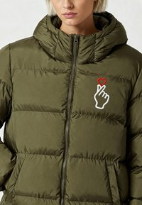 Veste matelassée vert olive avec une fermeture éclair, une capuche et une main brodée avec un motif de cœur sur le côté gauche. Texture douce.