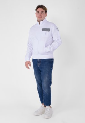 Giacca bianca con zip, tasca sul petto grigia, polsini e bordo a coste. Abbinata a jeans blu e sneakers bianche. Tessuto morbido, design casual.