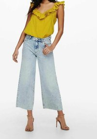 Gul volangstopp kombinerat med ljusblå vida jeans. Modellen bär nakna klacksandaler. Enkelt, avslappnat outfit med minimal detaljrikedom.