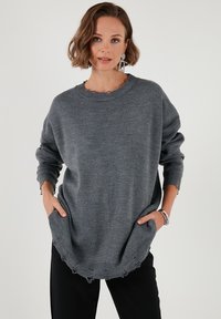 Szary oversize sweter z prążkowanym dołem i postrzępionymi brzegami. Posiada okrągły dekolt i długie rękawy. Kieszenie po bokach z przodu.