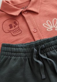 Kurzärmliges, korallenfarbenes Polo-Shirt mit einer Totenkopf-Stickerei und hellen Knöpfen, kombiniert mit dunkelgrauen, weichen Shorts mit elastischem Bund und Kordelzug.