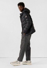 Zwarte donsjacket met capuchon, voorzien van een ritssluiting en een logoplaatje. Gecombineerd met grijze broek en sportieve schoenen in neutrale tinten.