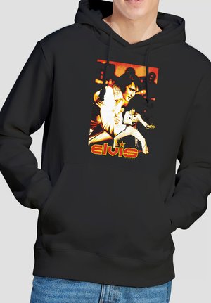 Person trägt schwarzen Hoodie mit orange-gelbem Elvis-Presley-Grafik, die ihn in einem weißen Overall zeigt, sowie dem Schriftzug "elvis".