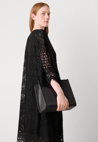 Black lace dress with intricate patterns and open weave, worn with a structured, black leather handbag featuring a textured finish.