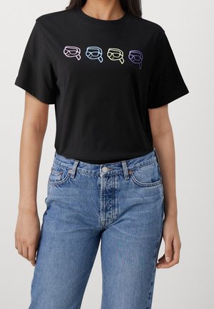 Kvinde iført sort T-shirt med fire farverige optrukne ansigter med solbriller over brystet, kombineret med blå jeans.