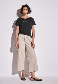 T-shirt en coton noir avec "FIND" imprimé en blanc, associé à un pantalon large beige clair et des chaussures noires en mesh à enfiler.