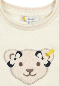 Cremfarbener Sweatshirt mit einem Bären-Applikation. Enthält gestickte Details, einschließlich einer Schleife am Ohr des Bären und einem gelben Akzentanhänger.