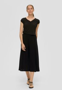 Zwart, kort-sleeve top met een V-hals en geplooide schouderdetails, gecombineerd met een vloeiende zwarte rok. Model draagt zwarte platte sandalen.