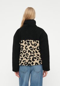 Svart og beige leopardmønstret fleecejakke med høy krage, teksturert stoff og lange ermer. Dongeribukser er synlige nederst.
