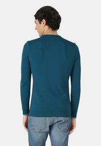 Uomo che indossa una camicia a maniche lunghe color turchese e jeans azzurri, visto da dietro su uno sfondo bianco uniforme.