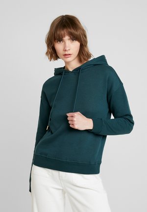 Teal cotton hoodie with a drawstring hood, ribbed cuffs, and hem. Model wearing it with cream-colored pants against a neutral background.