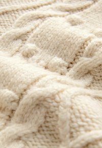 Cremefarvet strikket sweater med et tekstureret kabelmønster, der har tykt garn og et blødt, fyldigt udseende. Nærbillede fremhæver detaljer.