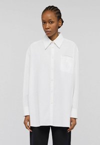 Donna con i capelli intrecciati, che indossa una camicia bianca oversize con colletto ampio e taschino frontale, abbinata a pantaloni neri.