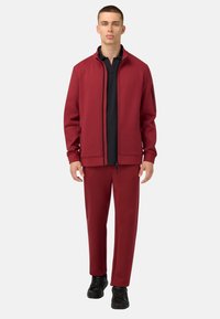 Rote Zip-Jacke mit schwarzen Akzenten, kombiniert mit passenden roten Hosen. Schwarz gepolsterte Shirt darunter. Das Modell steht vor einem weißen Hintergrund.