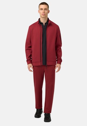 Rode zip-up jas met zwarte accenten, gecombineerd met bijpassende rode broek. Zwart poloshirt eronder. Model staat tegen een witte achtergrond.