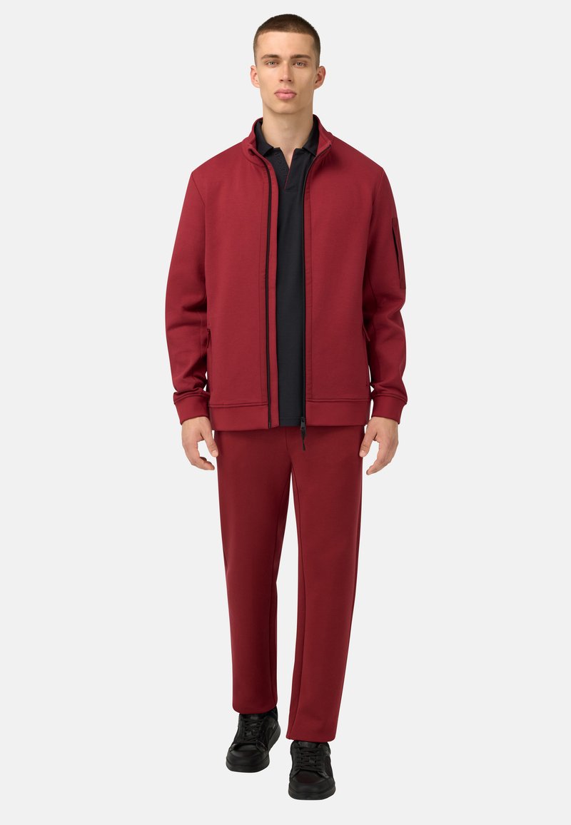 Rote Zip-Jacke mit schwarzen Akzenten, kombiniert mit passenden roten Hosen. Schwarz gepolsterte Shirt darunter. Das Modell steht vor einem weißen Hintergrund.