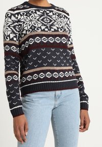 Pull tricoté avec une base marine ornée de motifs géométriques blancs et bordeaux. Manches longues, col rond, poignets et ourlet côtelés.
