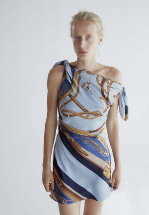 Mujer rubia con un vestido azul de un solo hombro con un cinturón marrón y dorado y un patrón de cadenas, de pie contra un fondo claro.