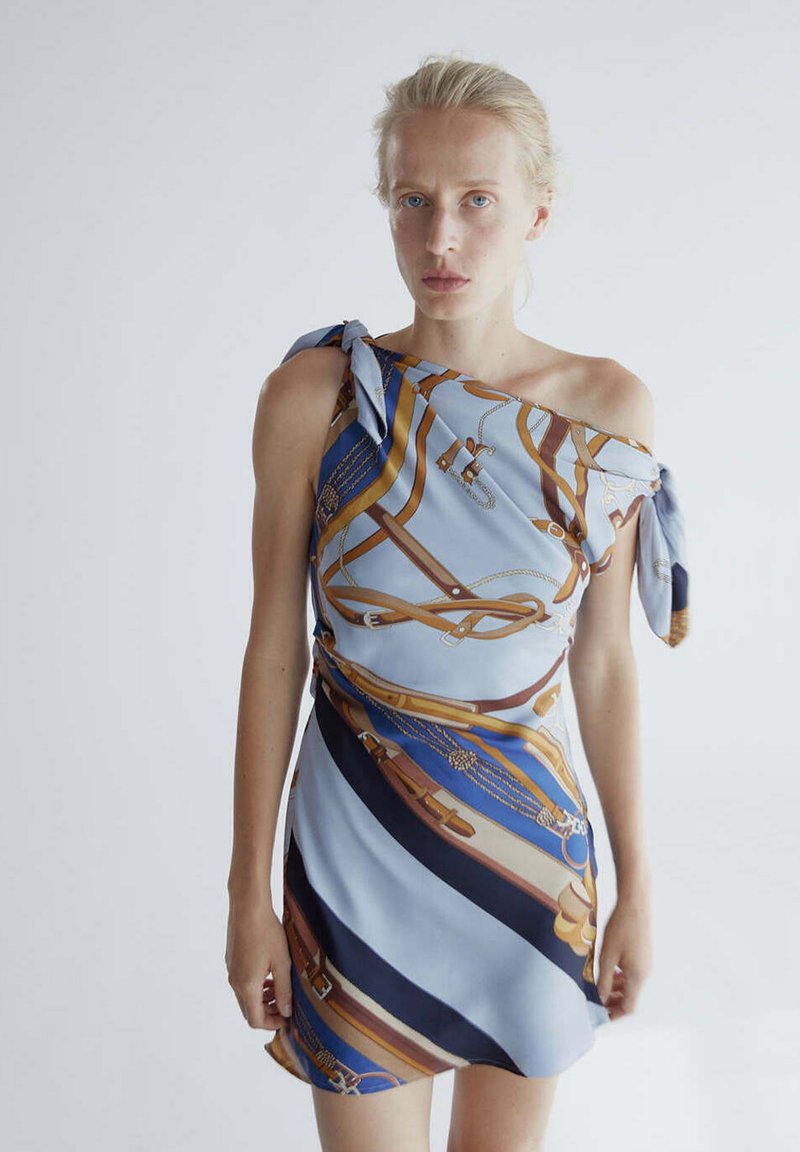 Femme blonde portant une robe bleue asymétrique avec une ceinture marron et or et un motif chaîne, debout sur un fond clair.