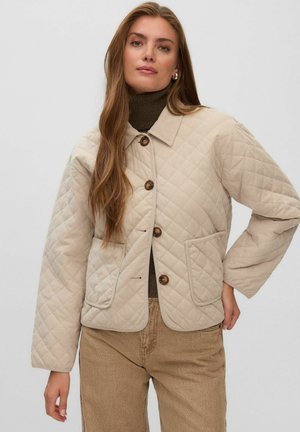 Femme aux longs cheveux portant une veste matelassée beige par-dessus un col roulé marron et un pantalon marron clair, debout, une main sur la hanche.