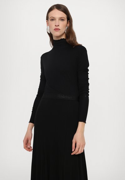 Tricou negru cu mânecă lungă și guler înalt, cu o siluetă ajustată și detalii subtile cu logo la talie, asortat cu o fustă lungă neagră și fluidă.